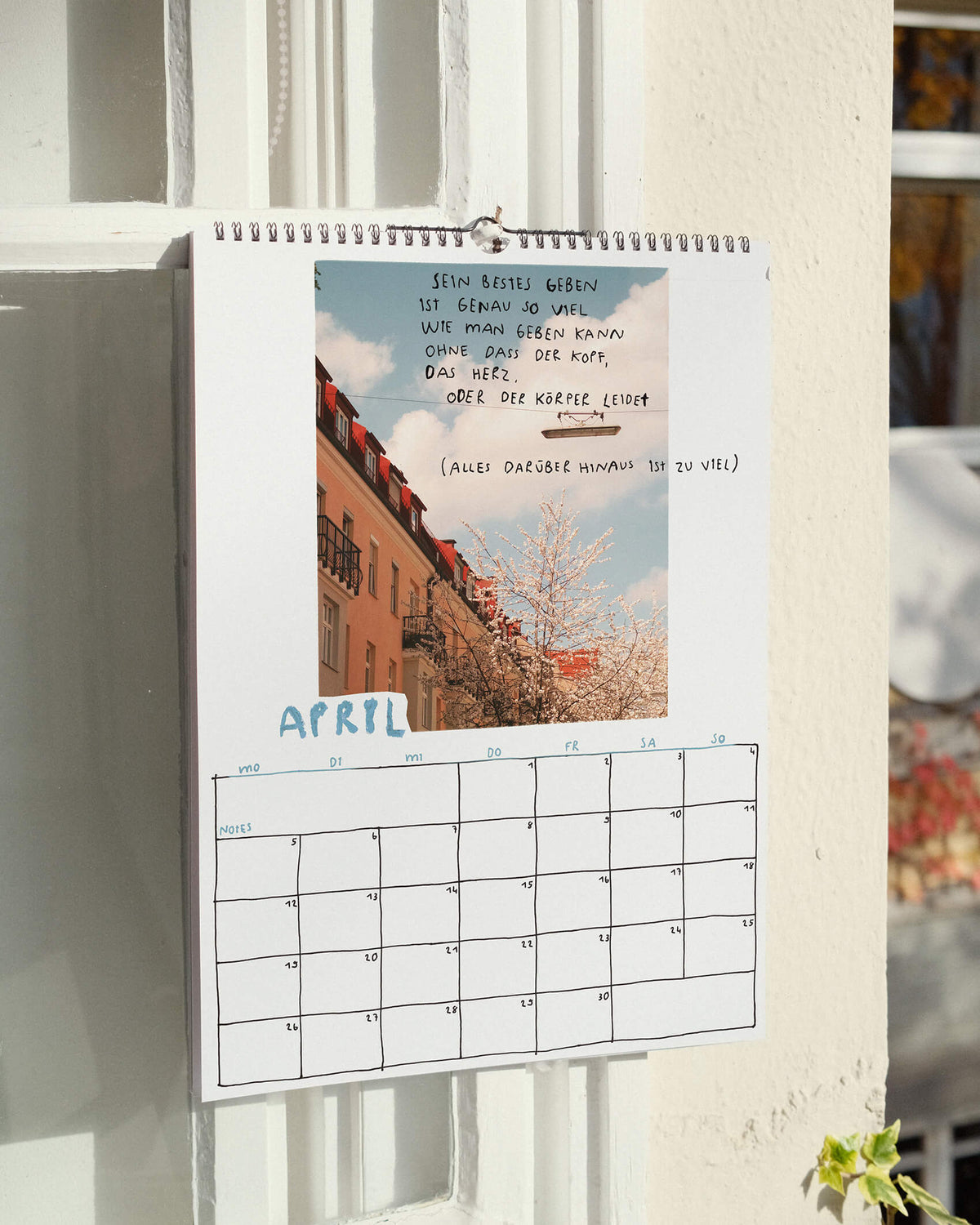 Aprilblatt des Wandkalenders 2027 von Angela Doe: blühender Baum vor Häusern und blauem Himmel mit einem handgeschriebenen Spruch über Selbstfürsorge und eigene Grenzen.