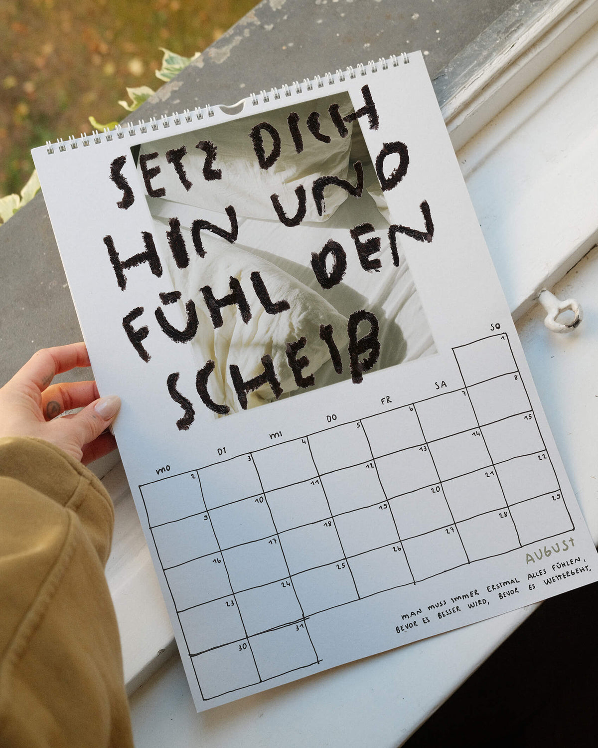 Augustblatt des Wandkalenders 2027 von Angela Doe: helle Bettwäsche im Sonnenlicht mit dem handgeschriebenen Spruch „Setz dich hin und fühl den Scheiß“ über das Zulassen von Gefühlen.