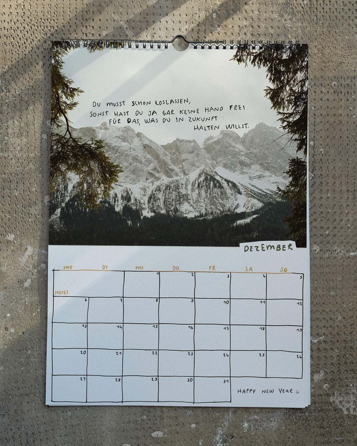 Dezemberblatt des Wandkalenders 2027 von Angela Doe: verschneite Berge zwischen Tannenzweigen mit einem handgeschriebenen Spruch über das Loslassen und Offenheit für die Zukunft.
