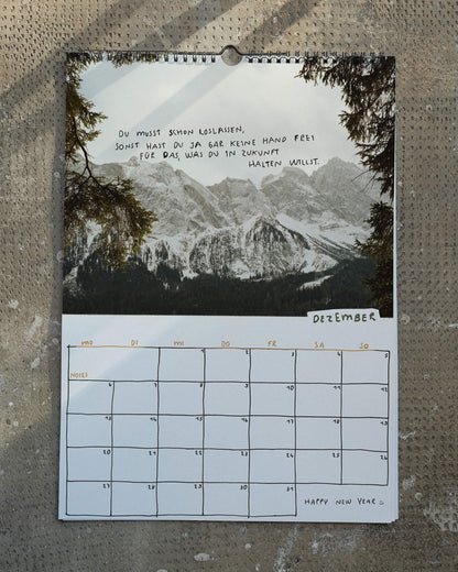Dezemberblatt des Wandkalenders 2027 von Angela Doe: verschneite Berge zwischen Tannenzweigen mit einem handgeschriebenen Spruch über das Loslassen und Offenheit für die Zukunft.