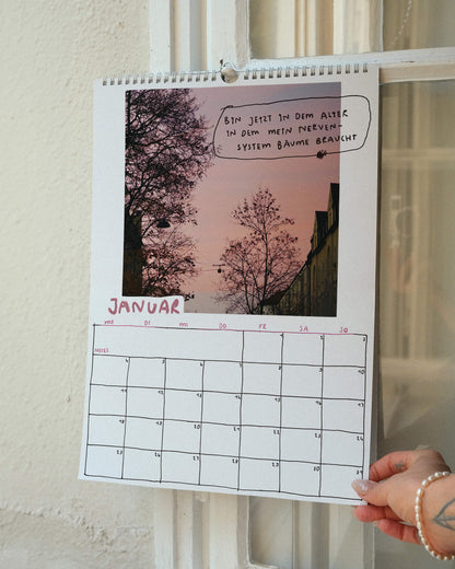 Januarblatt des Sprüchekalenders 2027 von Angela Doe: Bäume vor rosa Abendhimmel mit einem handgeschriebenen Spruch über das Bedürfnis nach Natur und Ruhe.