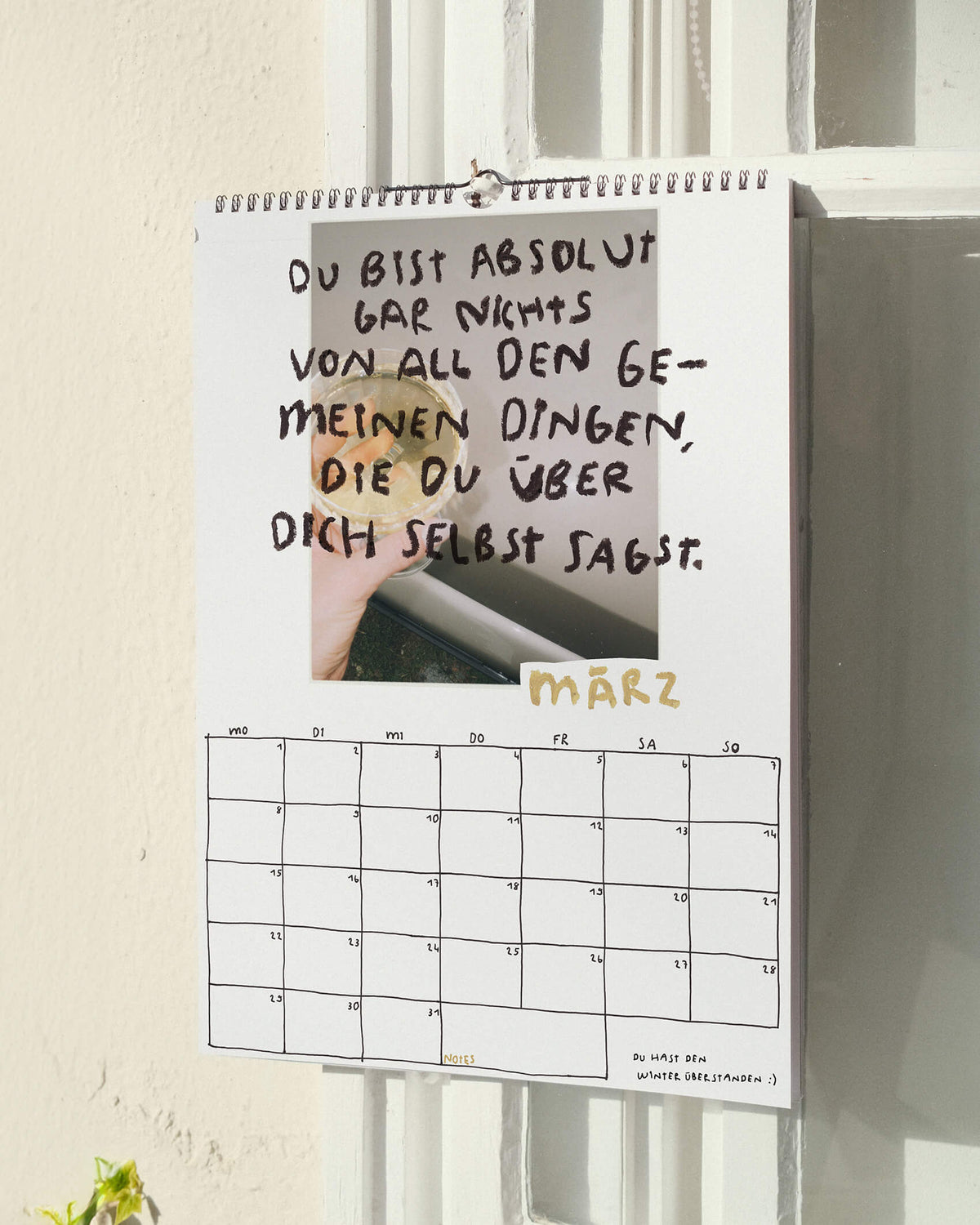 Märzblatt des Sprüchekalenders 2027 von Angela Doe: Hand mit einem Glas im Sonnenlicht und dem Spruch „Du bist absolut gar nichts von all den gemeinen Dingen, die du über dich selbst sagst.“