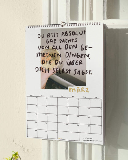 Märzblatt des Sprüchekalenders 2027 von Angela Doe: Hand mit einem Glas im Sonnenlicht und dem Spruch „Du bist absolut gar nichts von all den gemeinen Dingen, die du über dich selbst sagst.“